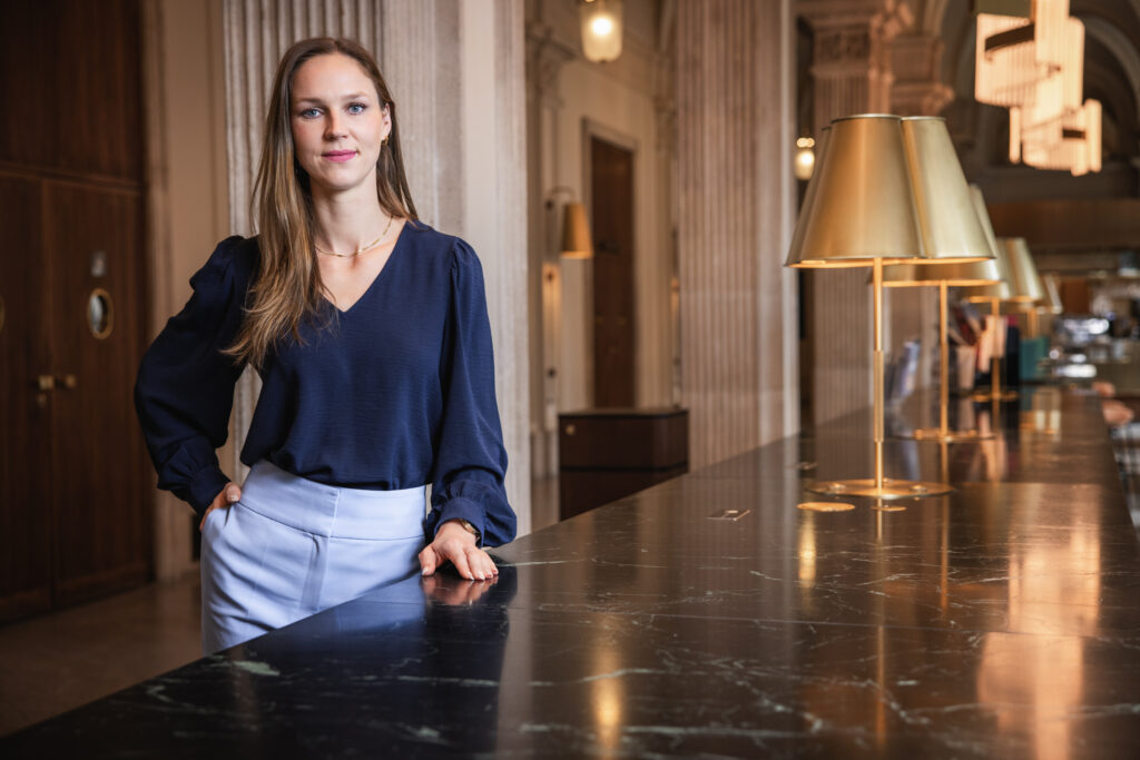 Eine Frau mit langen braunen Haaren steht an einem schwarzen Marmortresen in einem eleganten, hohen Raum mit Säulen und goldenen Lampen.