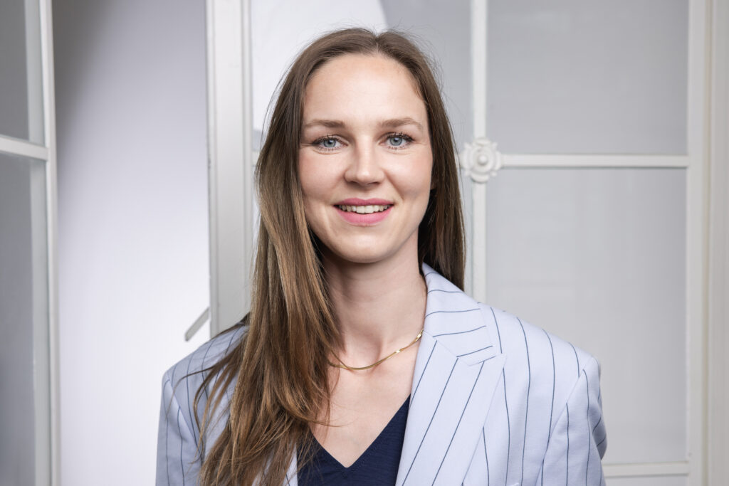 Junge Frau mit langen braunen Haaren lächelt, trägt einen hellblauen Blazer mit Streifen und steht vor einem Fenster.